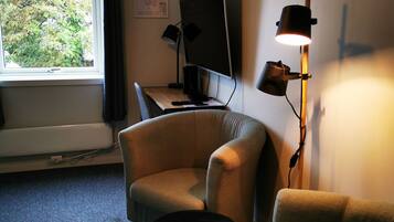 A room with a desk, two armchairs, a small round table, a standing lamp, and a window with a view of trees.