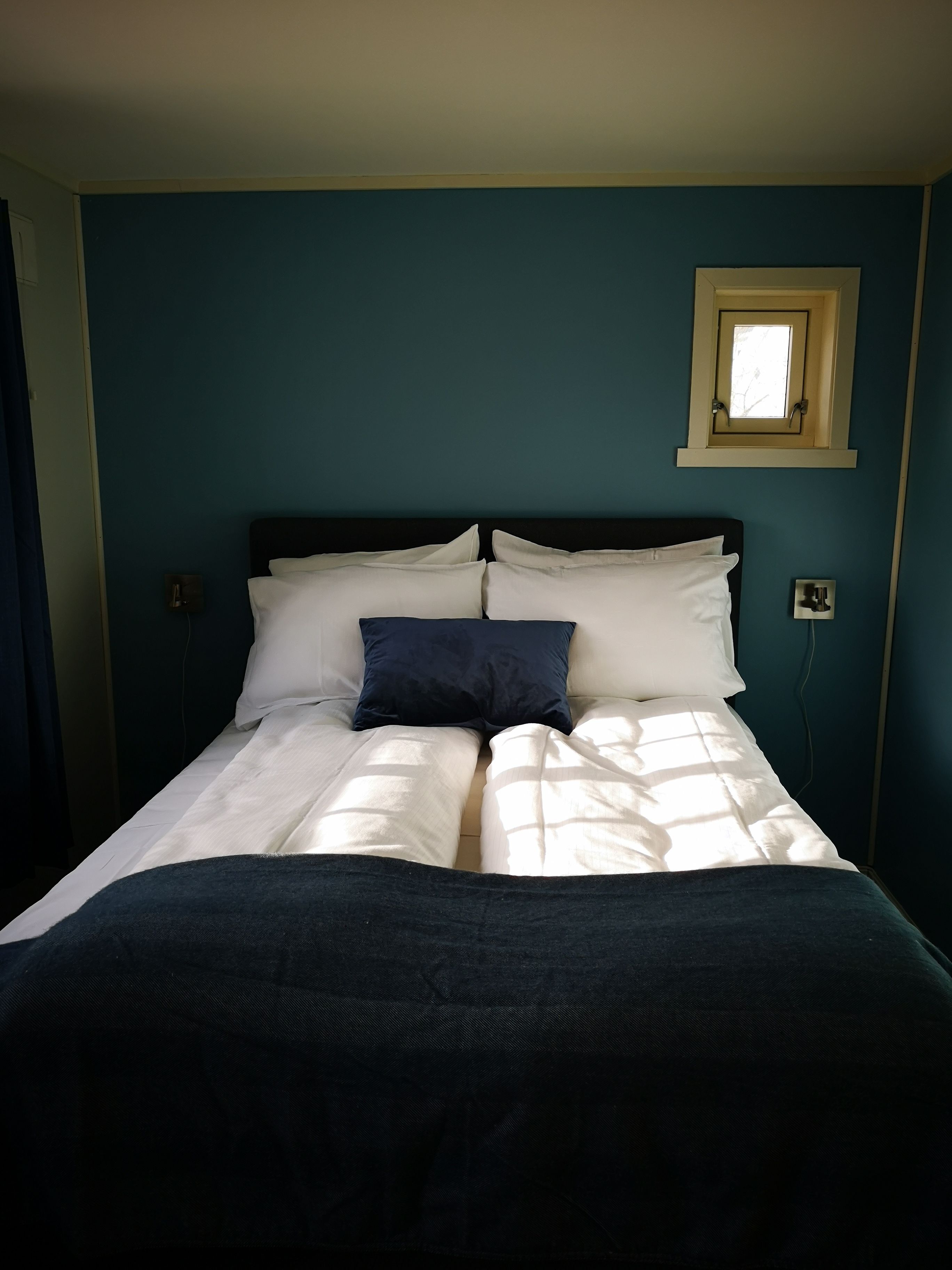 Uma cama bem arrumada com roupa de cama branca e azul escura, uma pequena janela acima da cabeceira e uma tomada na parede.
