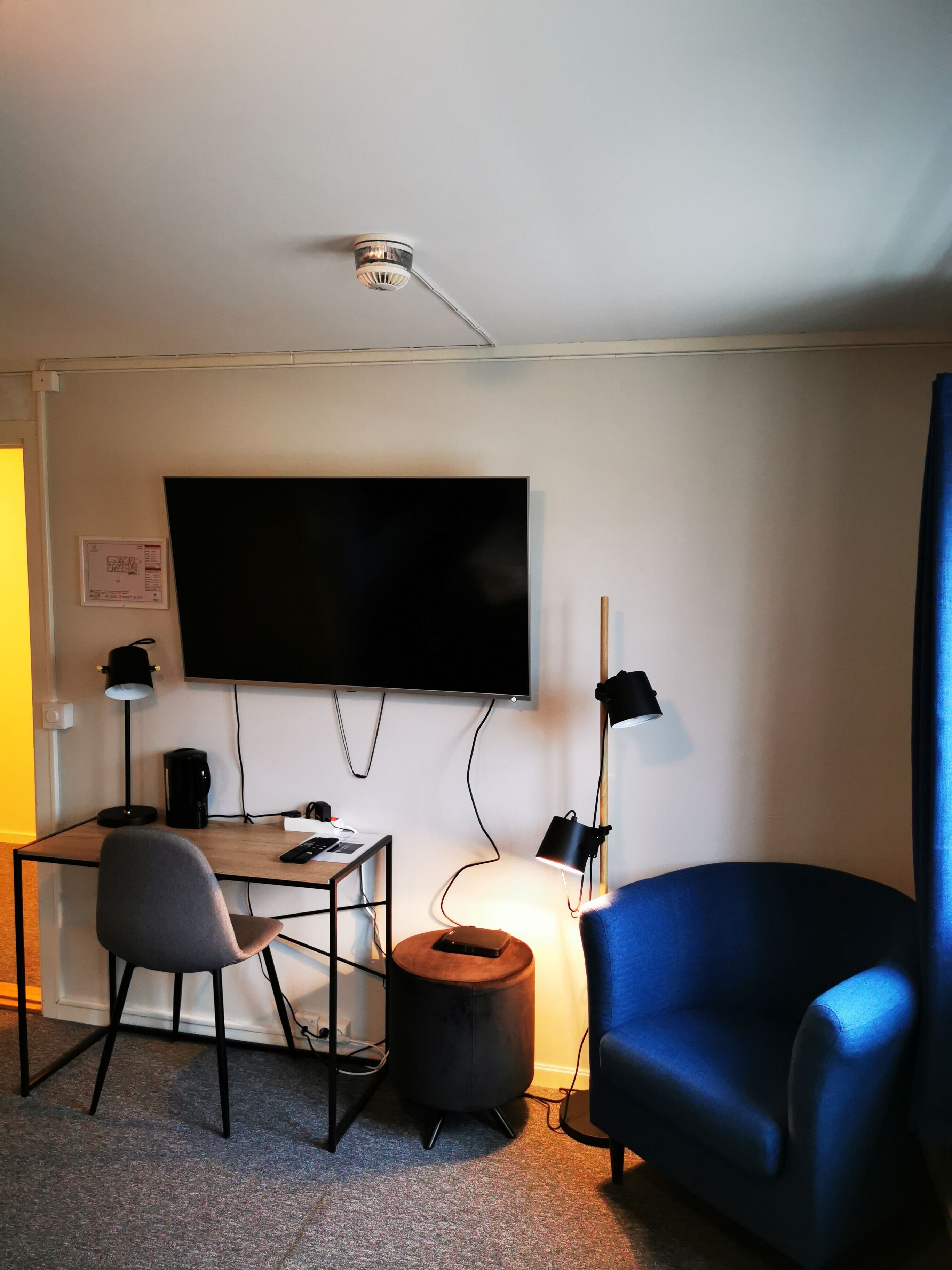 Una habitación con una televisión de pantalla plana montada en la pared, un escritorio con una silla, un sillón azul, una pequeña mesa redonda y dos lámparas de escritorio.
