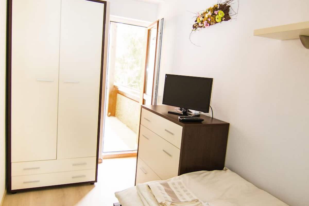 A bedroom with a bed, a desk with a computer, a wardrobe, and a window with a view of the outside.
