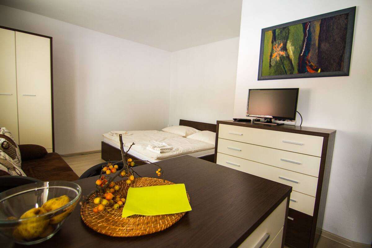 A hotel room with a bed, a TV on a stand, a desk with a basket of fruit and a yellow note, and a painting on the wall.