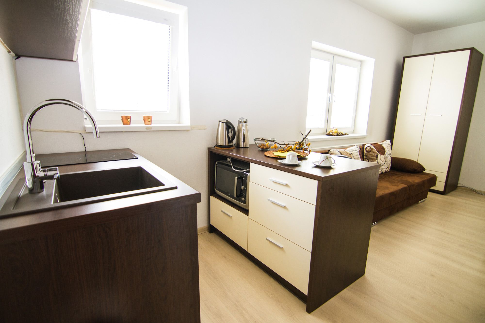 A modern kitchen with a sink, stove, and microwave. There is a window with a view of the outside. A small island with a kettle and a bowl of fruit is also visible.