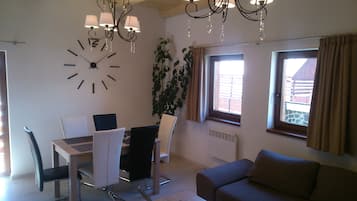 Un coin repas avec une table et des chaises, une grande horloge murale, un lustre, une plante en pot et une fenêtre avec des rideaux.