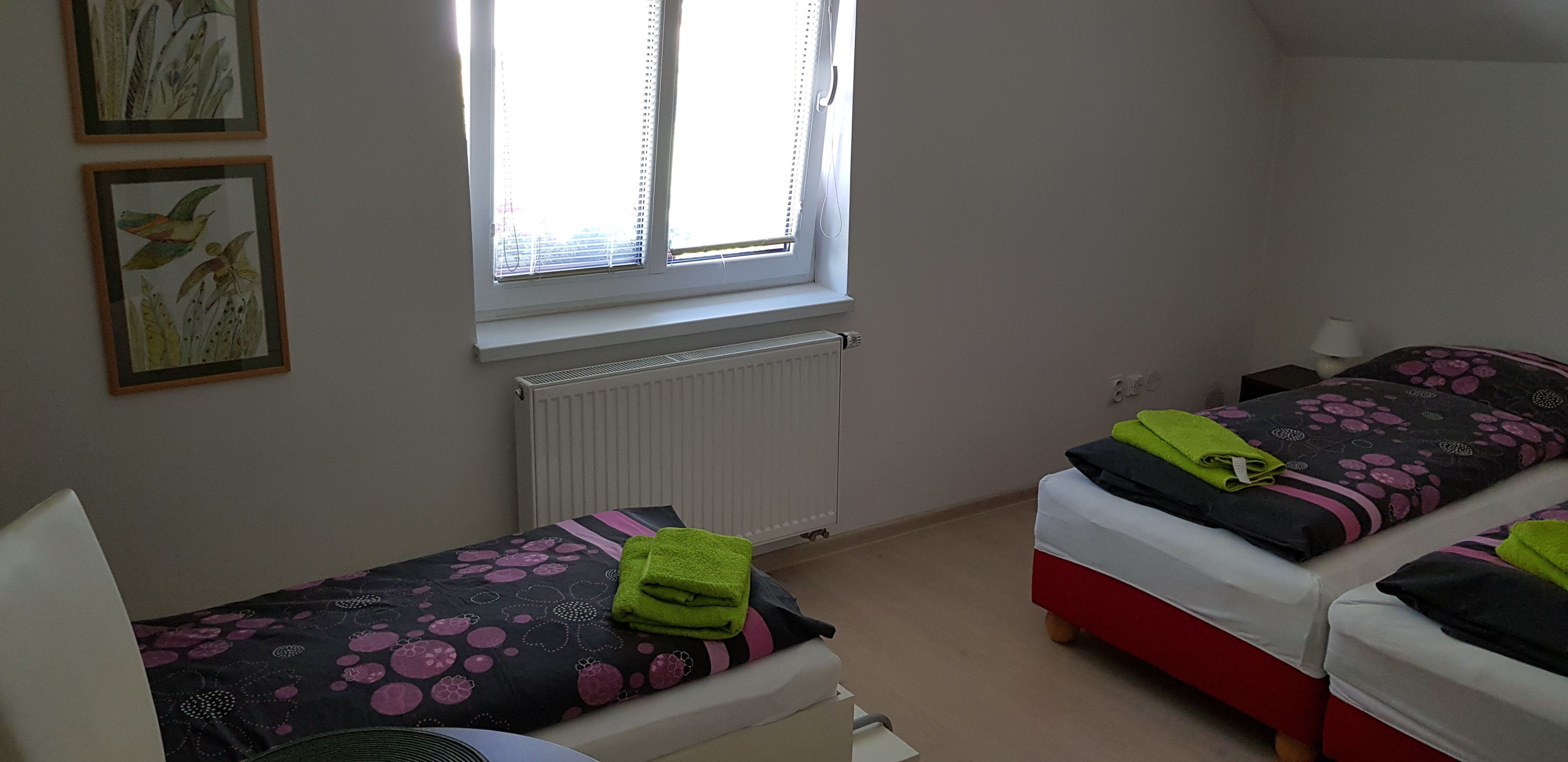 Une petite pièce propre, avec deux lits, un radiateur, une fenêtre et des œuvres d’art encadrées accrochées au mur.