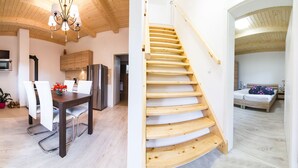 Una cocina moderna con armarios de madera, un refrigerador de acero inoxidable y un comedor con sillas blancas. Una escalera de madera lleva a un dormitorio con una cama y una mesita de noche.