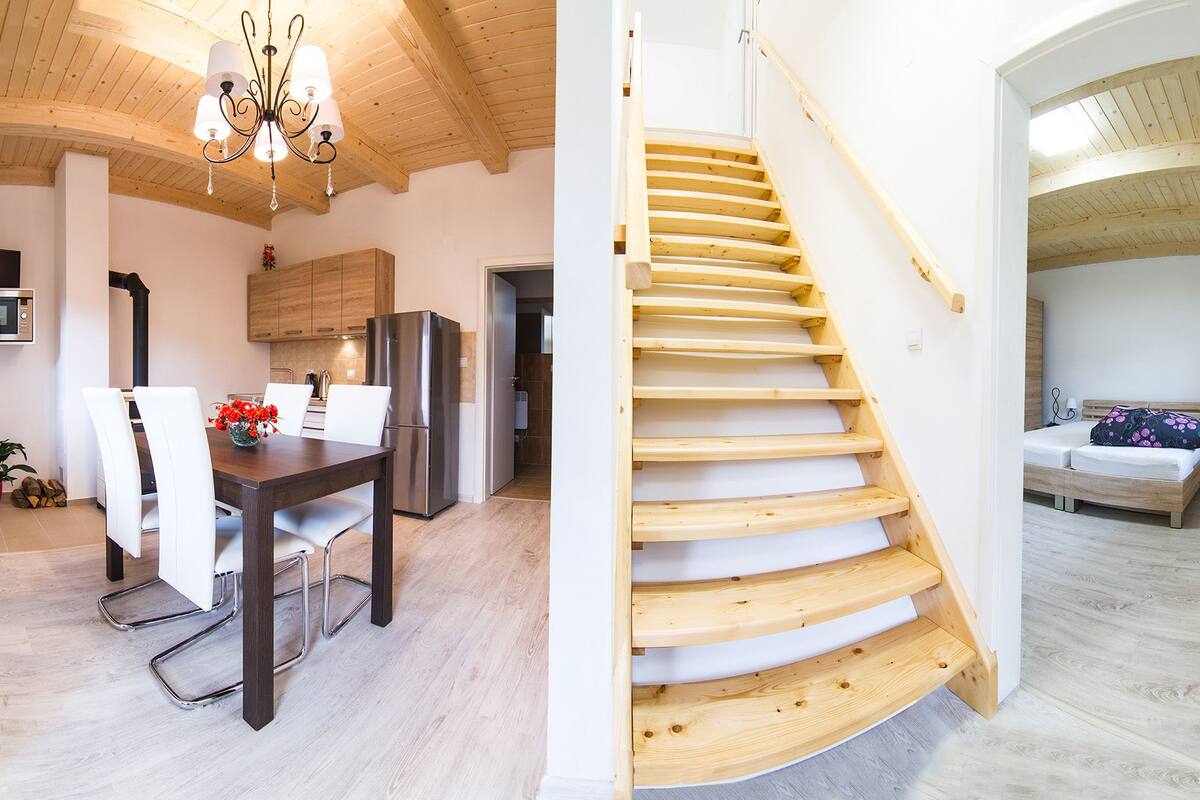 A modern kitchen with wooden cabinets, a stainless steel refrigerator, and a dining area with white chairs. A wooden staircase leads to a bedroom with a bed and a nightstand.
