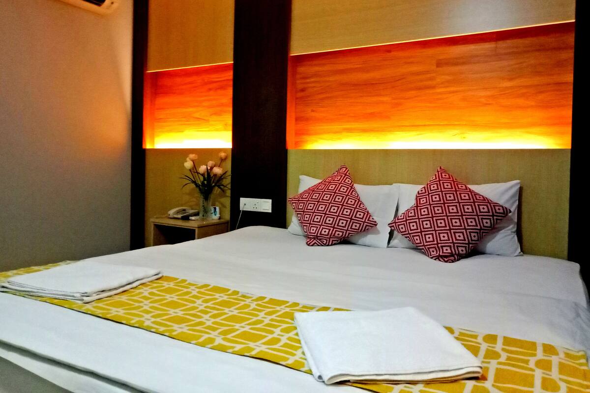 A hotel room with a bed, a nightstand, a vase with flowers, and a wall-mounted air conditioner.