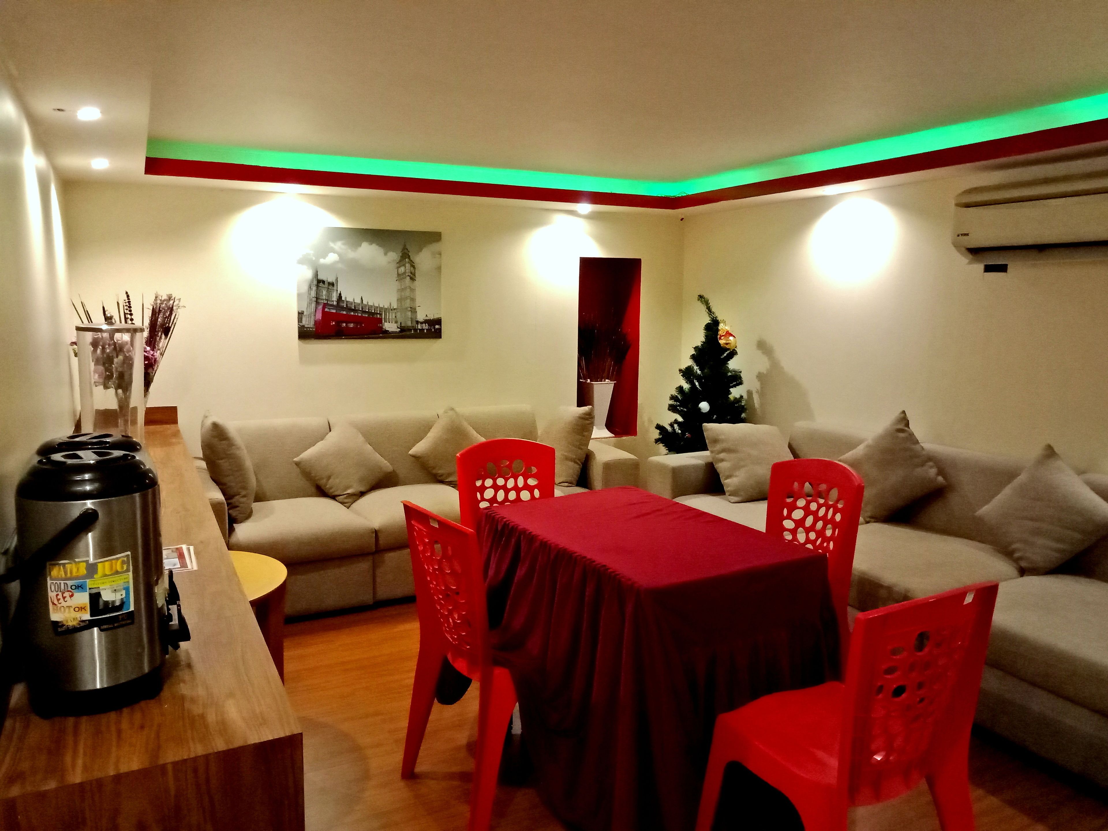 Una sala de estar con un sofá, una mesa de comedor con sillas rojas, una cafetera, un pequeño árbol de Navidad y un cuadro en la pared.