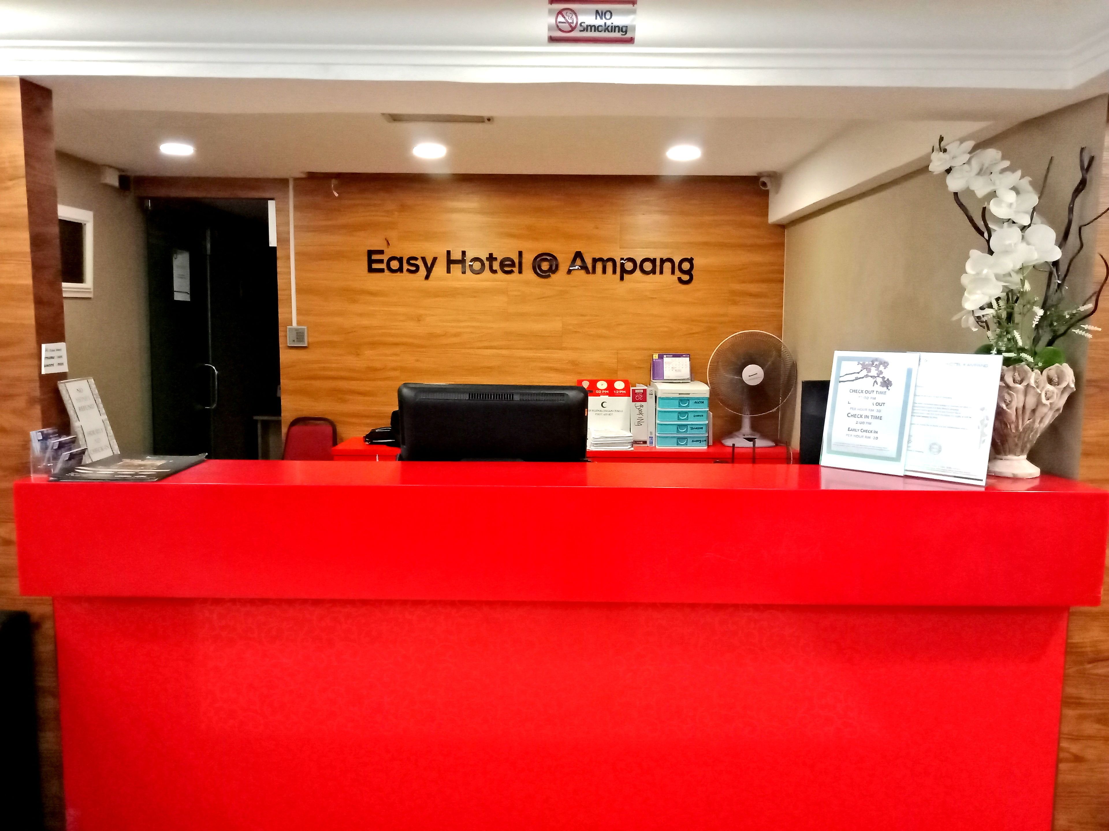 Una recepción de hotel con un mostrador rojo, una pared de madera con el nombre del hotel, un ventilador y un florero con flores.