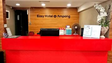 Una recepción de hotel con un mostrador rojo, una pared de madera con el nombre del hotel, un ventilador y un florero con flores.