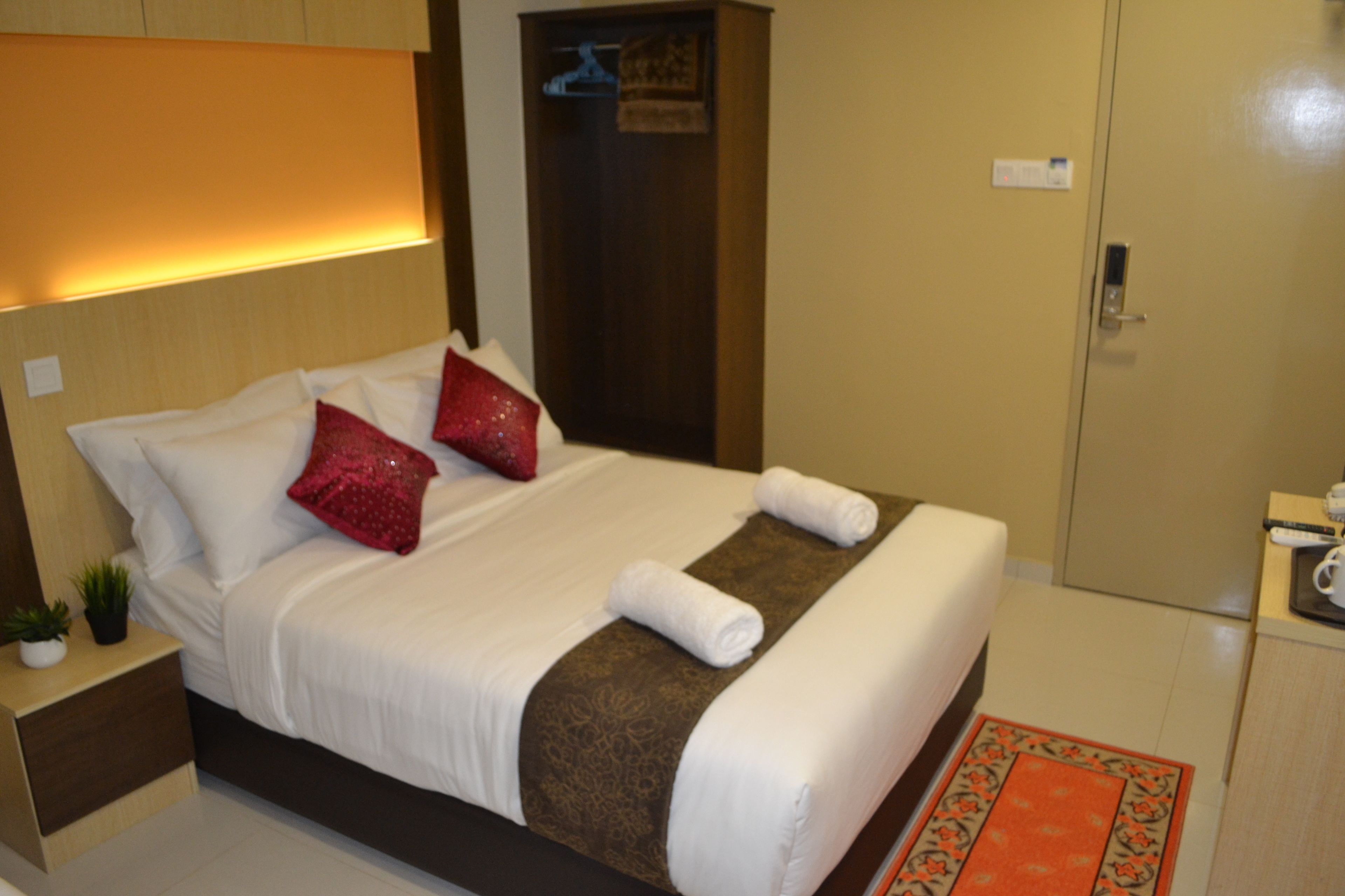 A neatly made bed with white linens, two red pillows, and a brown decorative runner. There is a small plant on the nightstand beside the bed.