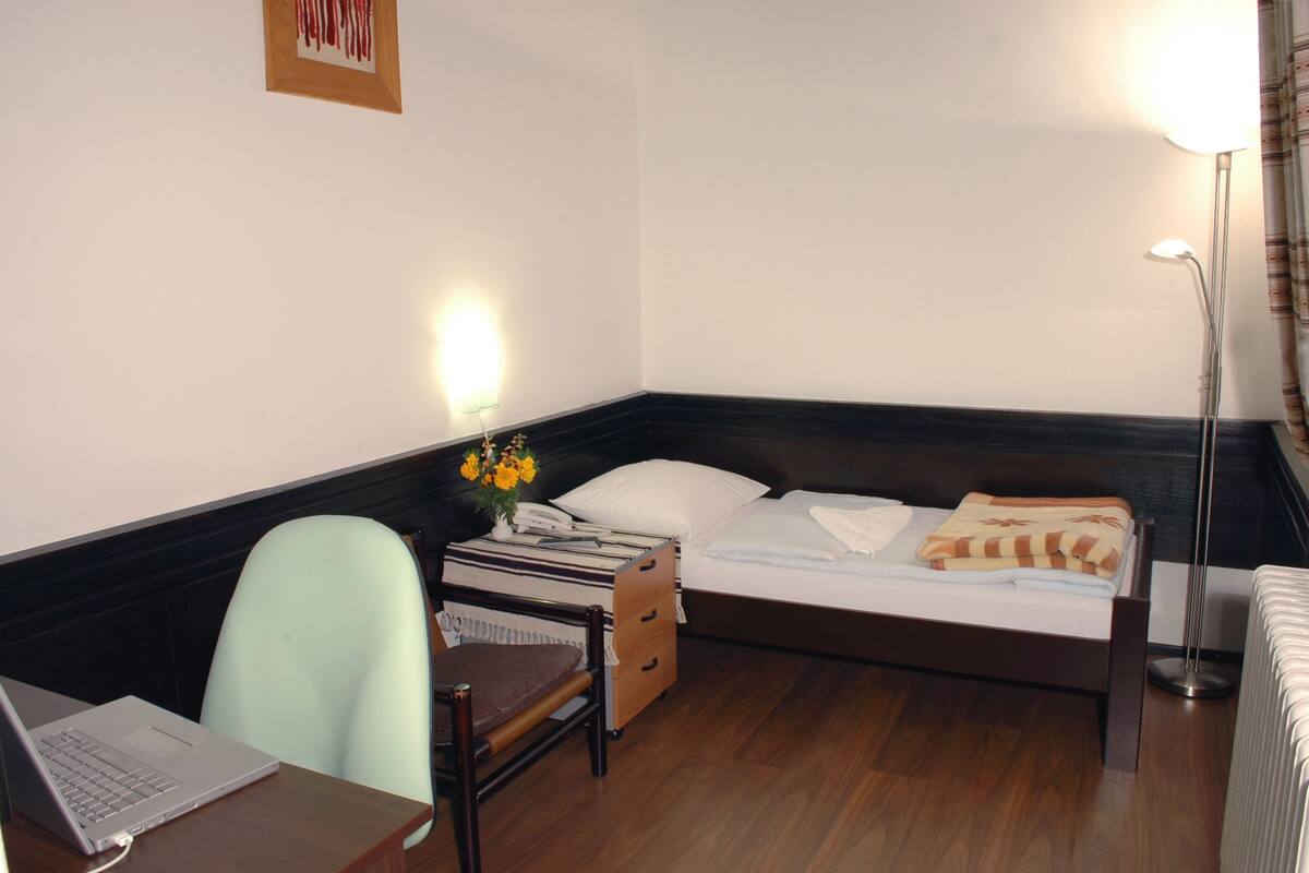 Een hotelkamer met een bed, een bureau met een laptop, een stoel, een nachtkastje met een bloemenvaas en een vloerlamp.