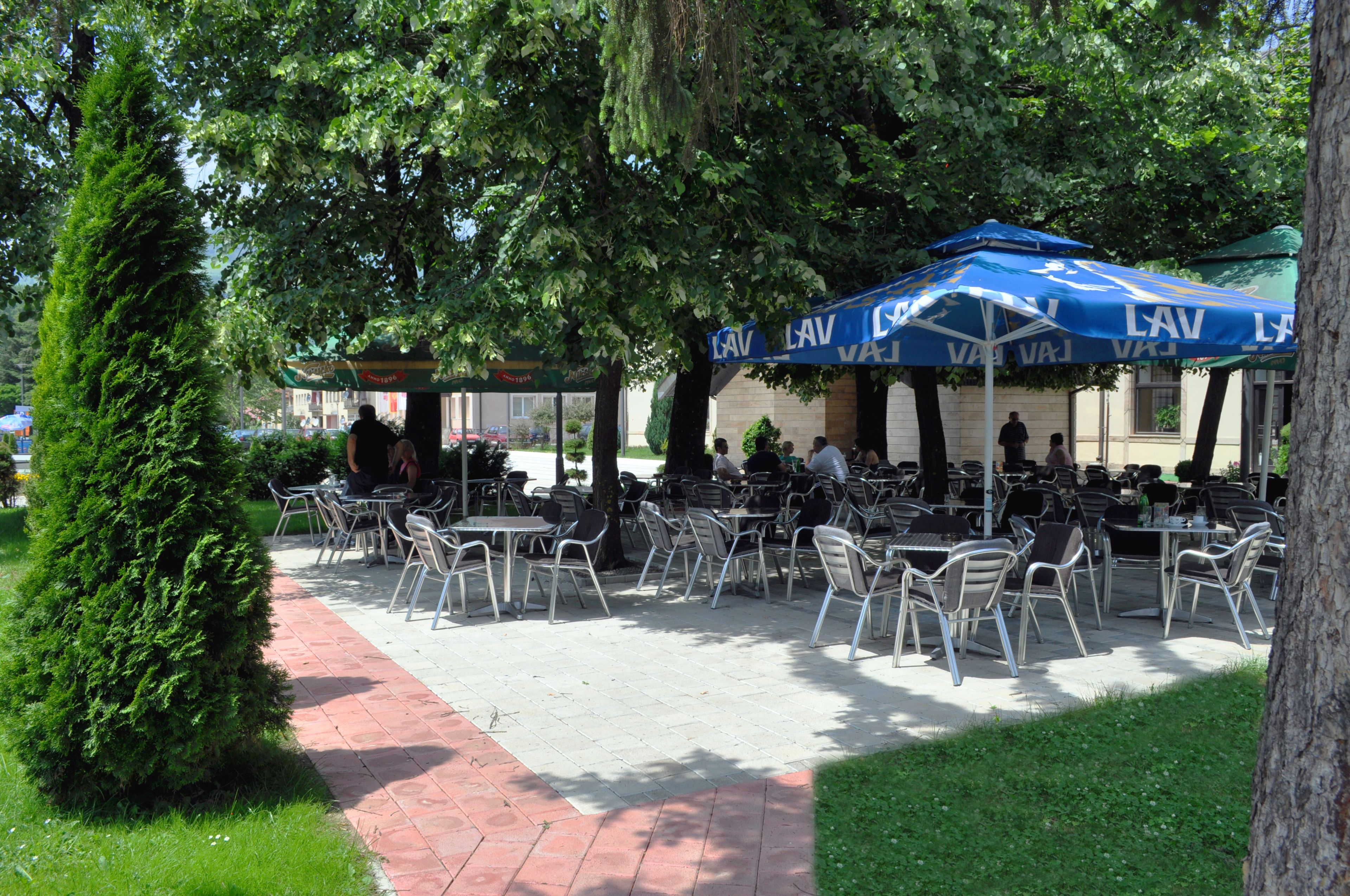 Un café al aire libre con mesas y sillas bajo sombrillas azules, rodeado de árboles.