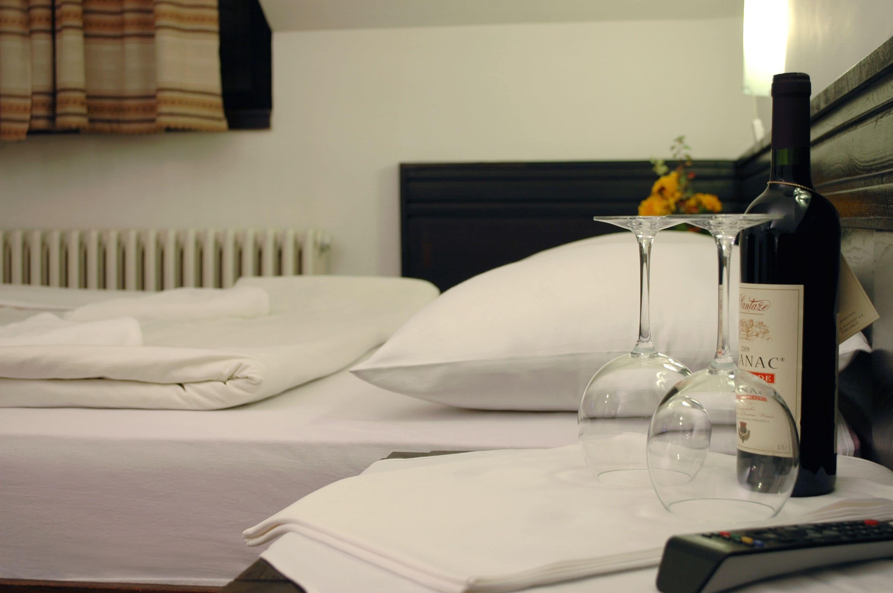 A neatly made bed with white linens, a bottle of wine, two wine glasses, and a remote control on a bedside table.
