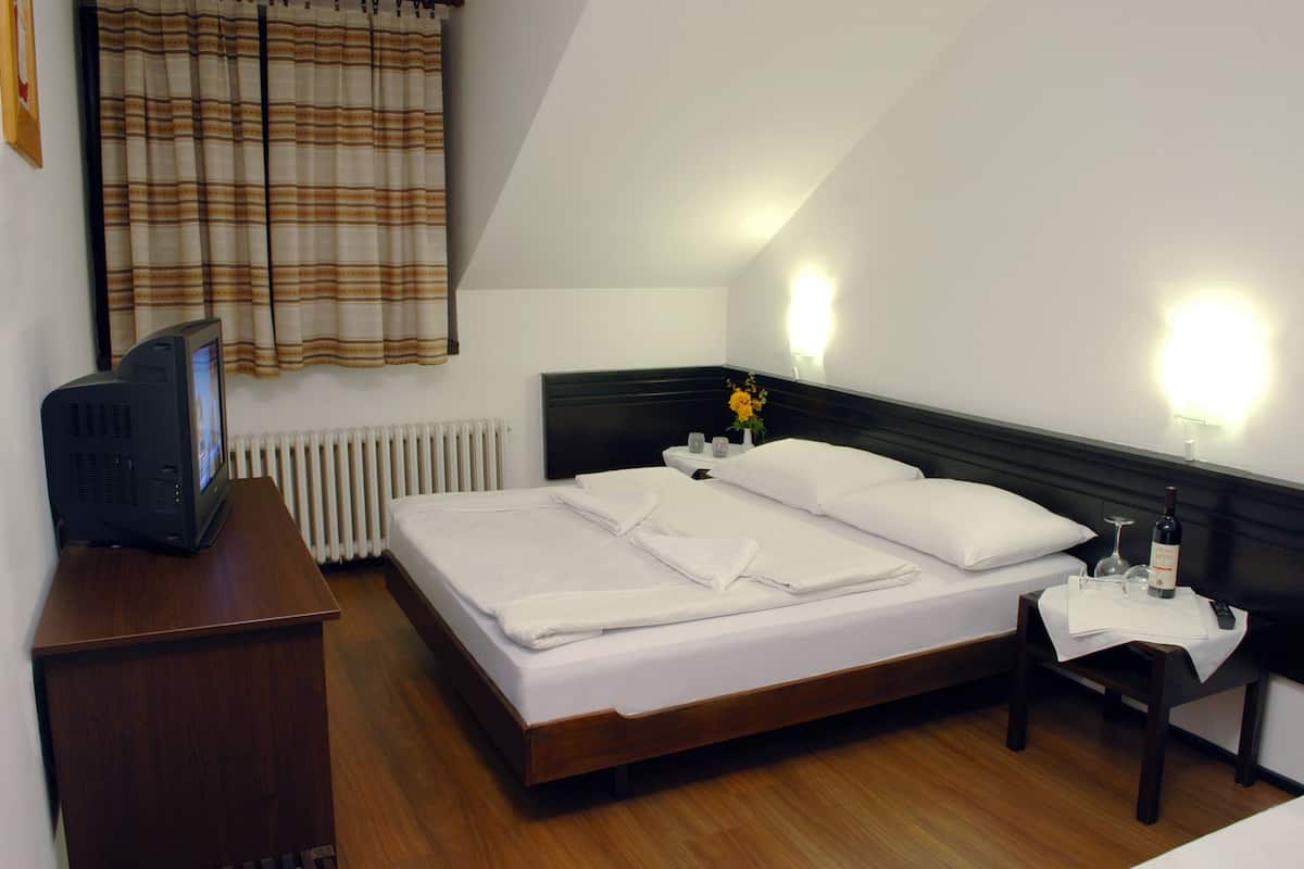 Een netjes opgemaakt bed met witte lakens, een nachtkastje met een fles en glazen, en een televisie op een houten plank.