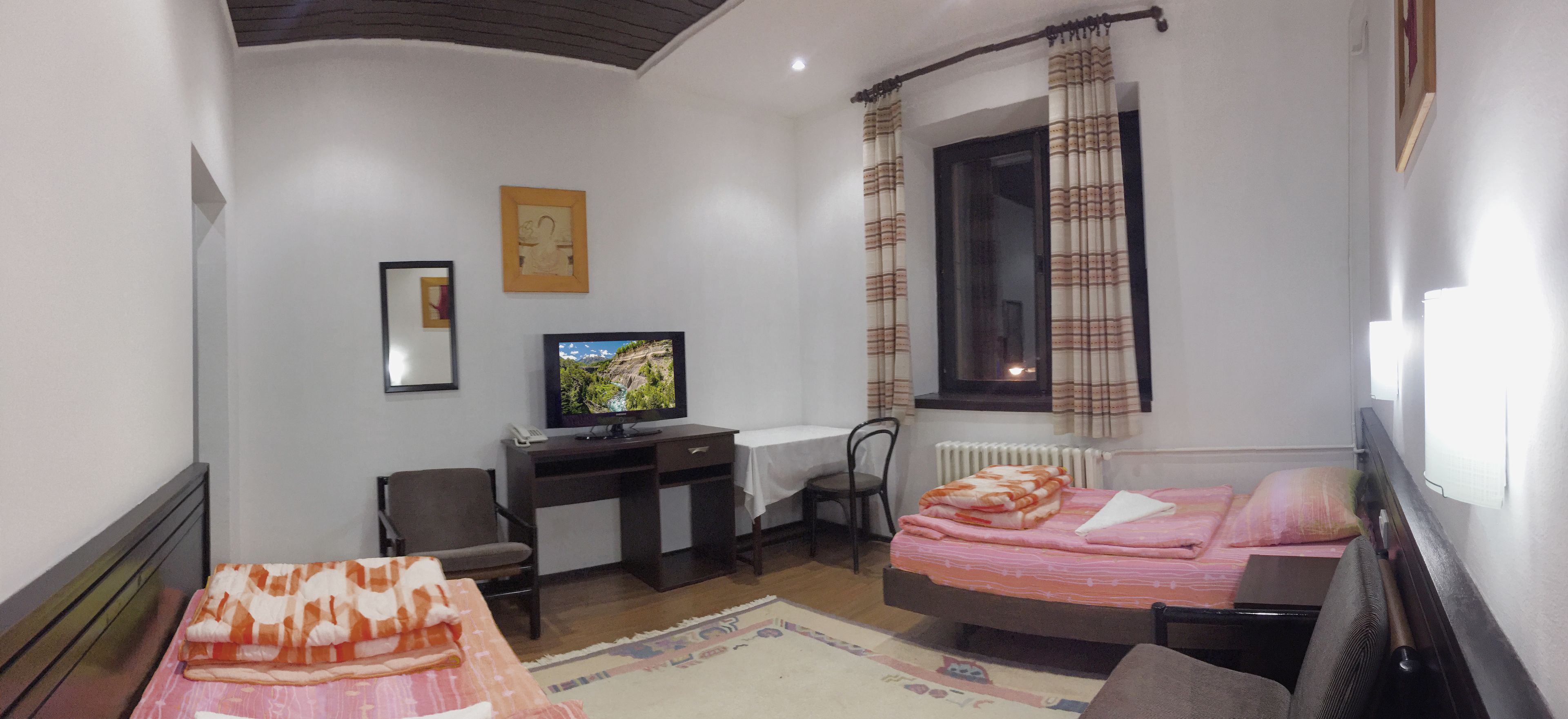 Een kleine, goed verlichte kamer met een bed, een bureau met een computer, een stoel en een raam met gordijnen.