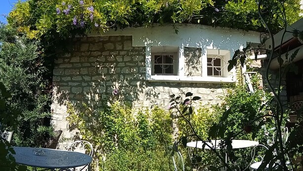 A stone building with three windows, surrounded by lush greenery and a small table with a glass top. - Hotel La Font de Lauro (Saumane-de-Vaucluse)