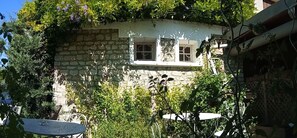 Un edificio de piedra con tres ventanas, rodeado de abundante vegetación y una pequeña mesa con superficie de vidrio.