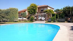 A swimming pool with a curved edge, surrounded by a concrete deck. There are two cars parked in the background, and a building with a tiled roof is visible behind the pool. - Hotel La Font de Lauro (Saumane-de-Vaucluse)