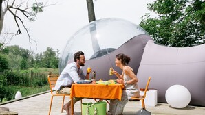 A couple dining at an outdoor table with a transparent dome overhead, surrounded by greenery and a wooden deck. - Les Aires en Scènes (Epagne-Epagnette)