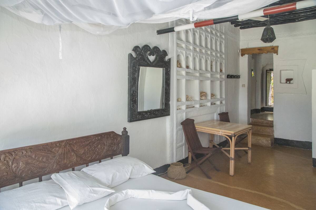 Ein Zimmer mit einem Bett, einer hölzernen Bank, einem Tisch und einem Stuhl. An der Wand hängt ein Spiegel und ein Regal mit Dekorationen.