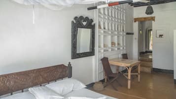 A room with a bed, a wooden bench, a table, and a chair. There is a mirror on the wall and a shelf with decorative items.