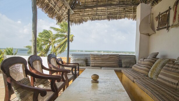 A beachside seating area with a table and chairs, a thatched roof, and a view of the sea. - Pwani Guest House (Lamu)