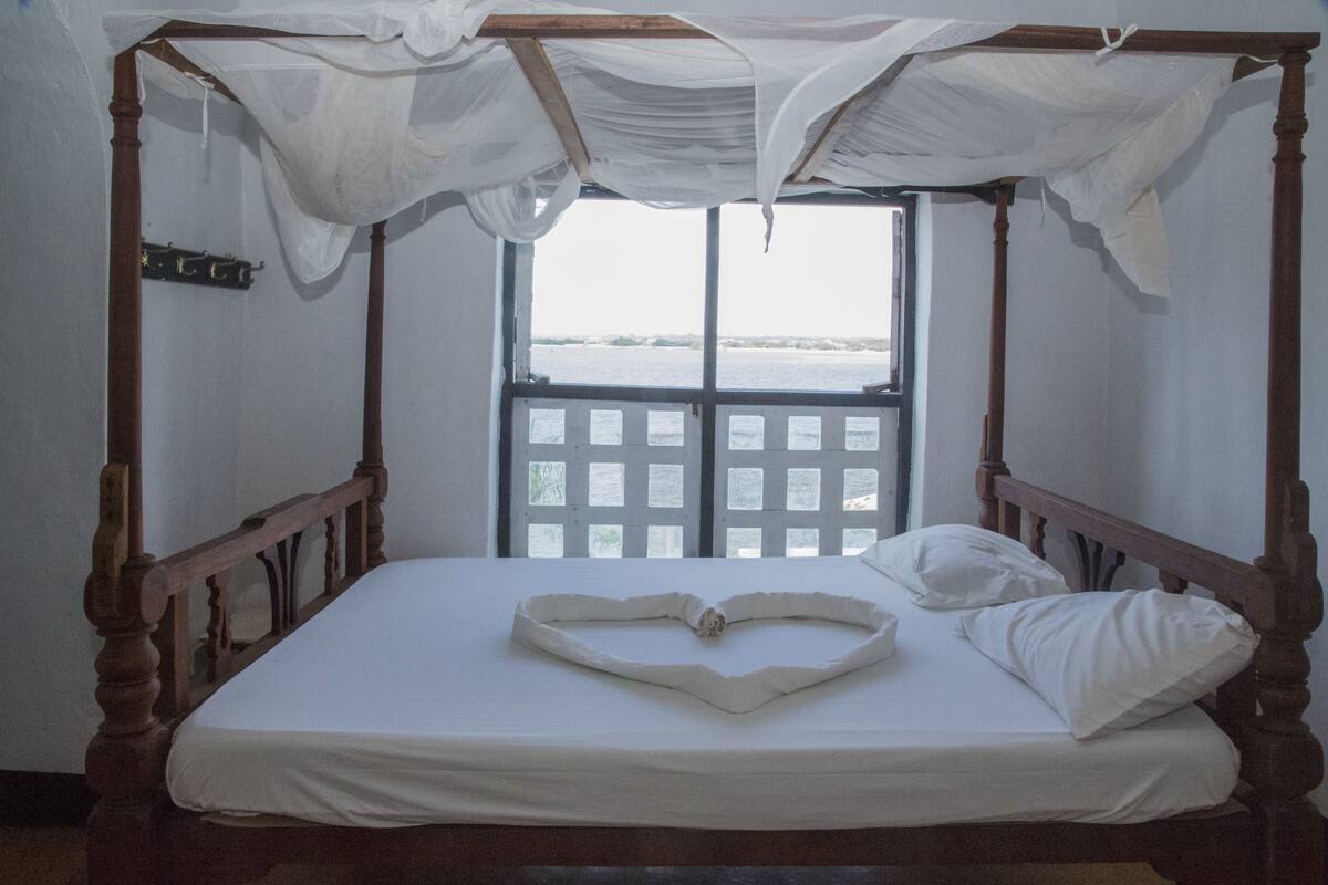 Ein Himmelbett mit einem herzförmigen Buch darauf, ein Fenster mit Blick auf das Meer und ein Holz-Kopfteil.
