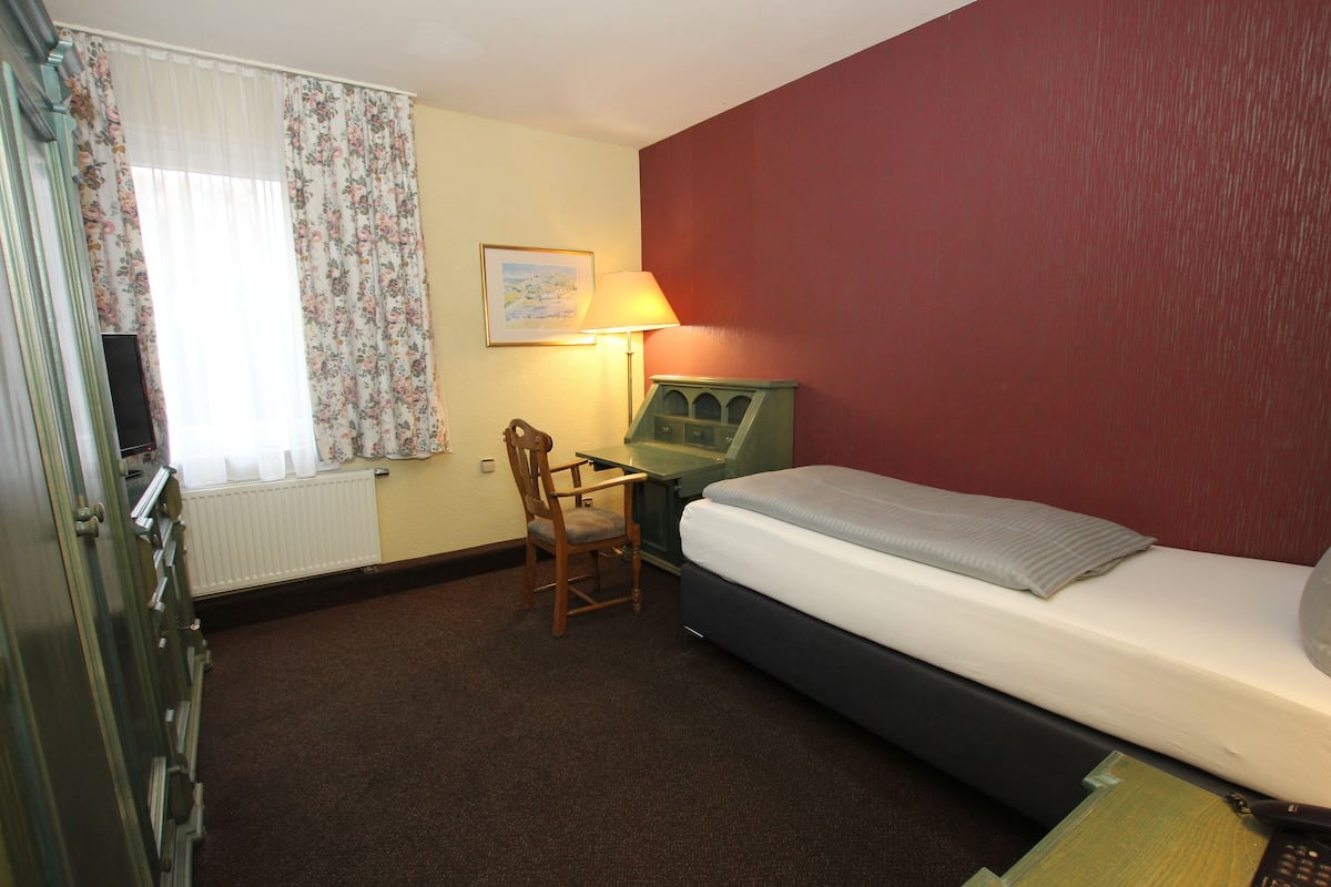 A hotel room with a bed, a desk, a chair, a lamp, and a radiator.