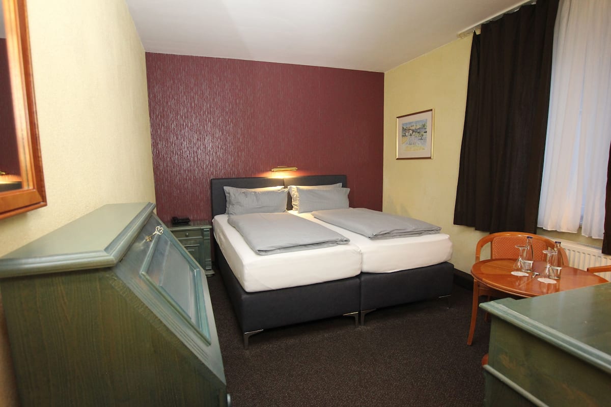 A hotel room with a double bed, a desk with a chair, a small table with a chair, and a window with curtains.