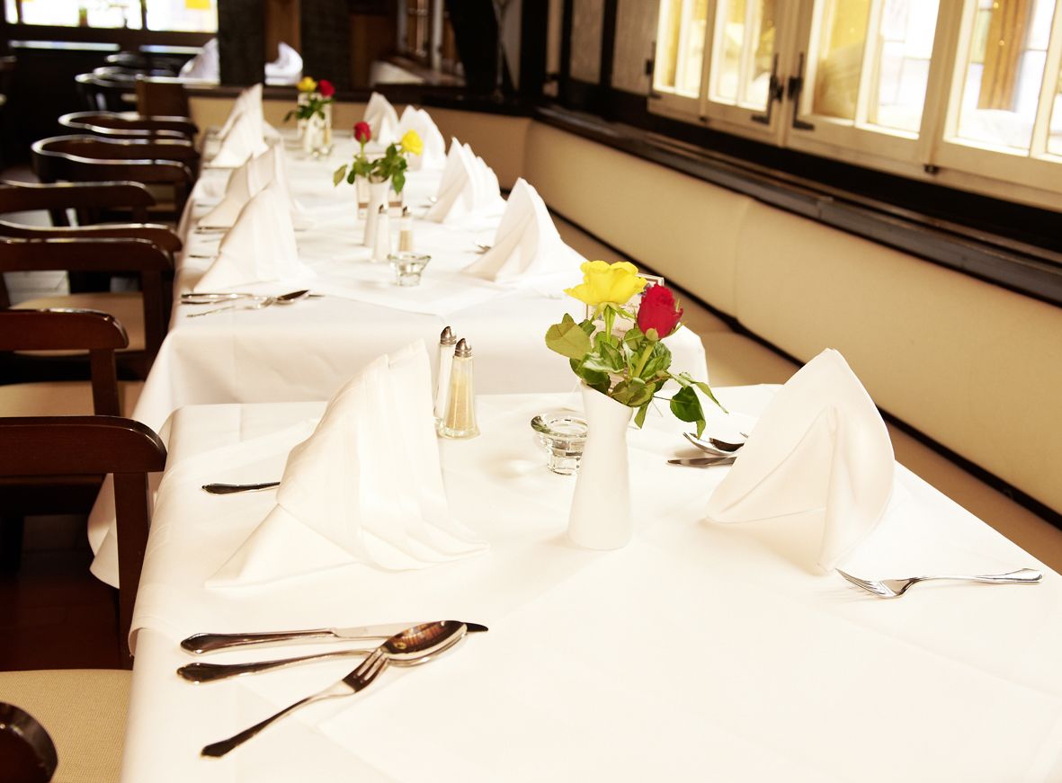 A dining area with tables set for service, each with a white tablecloth, silverware, and a vase with flowers.