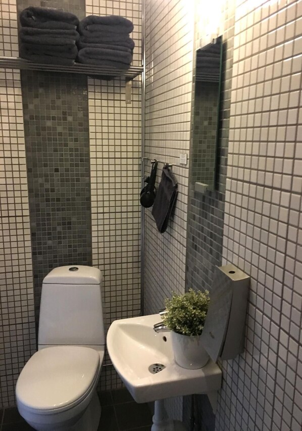 Un baño pequeño con inodoro, lavamanos y un espejo. Toallas apiladas en un estante sobre el inodoro.