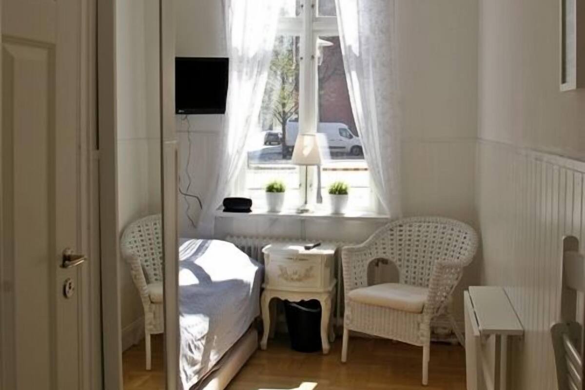 A small, well-lit bedroom with a bed, a chair, a small table, and a window with curtains.