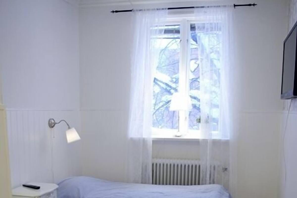 A small, single-room accommodation with two beds, a radiator, a window with sheer curtains, a bedside lamp, and a wooden table.