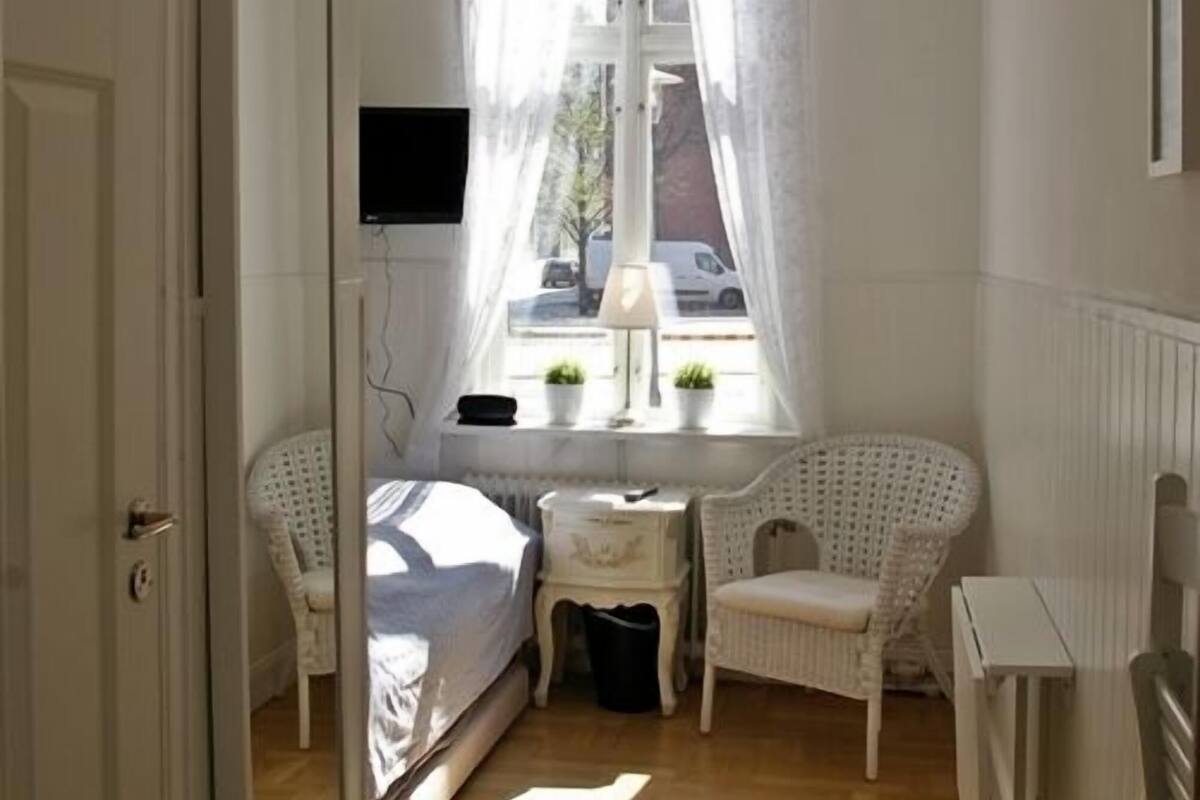 A small, bright room with a bed, two chairs, a small table, a mirror, and a window with curtains.
