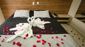A neatly made bed with a black and white patterned bedspread, two pillows, a pair of white towels arranged in a heart shape, and scattered red rose petals. - Pousada Chamamé (Bonito)