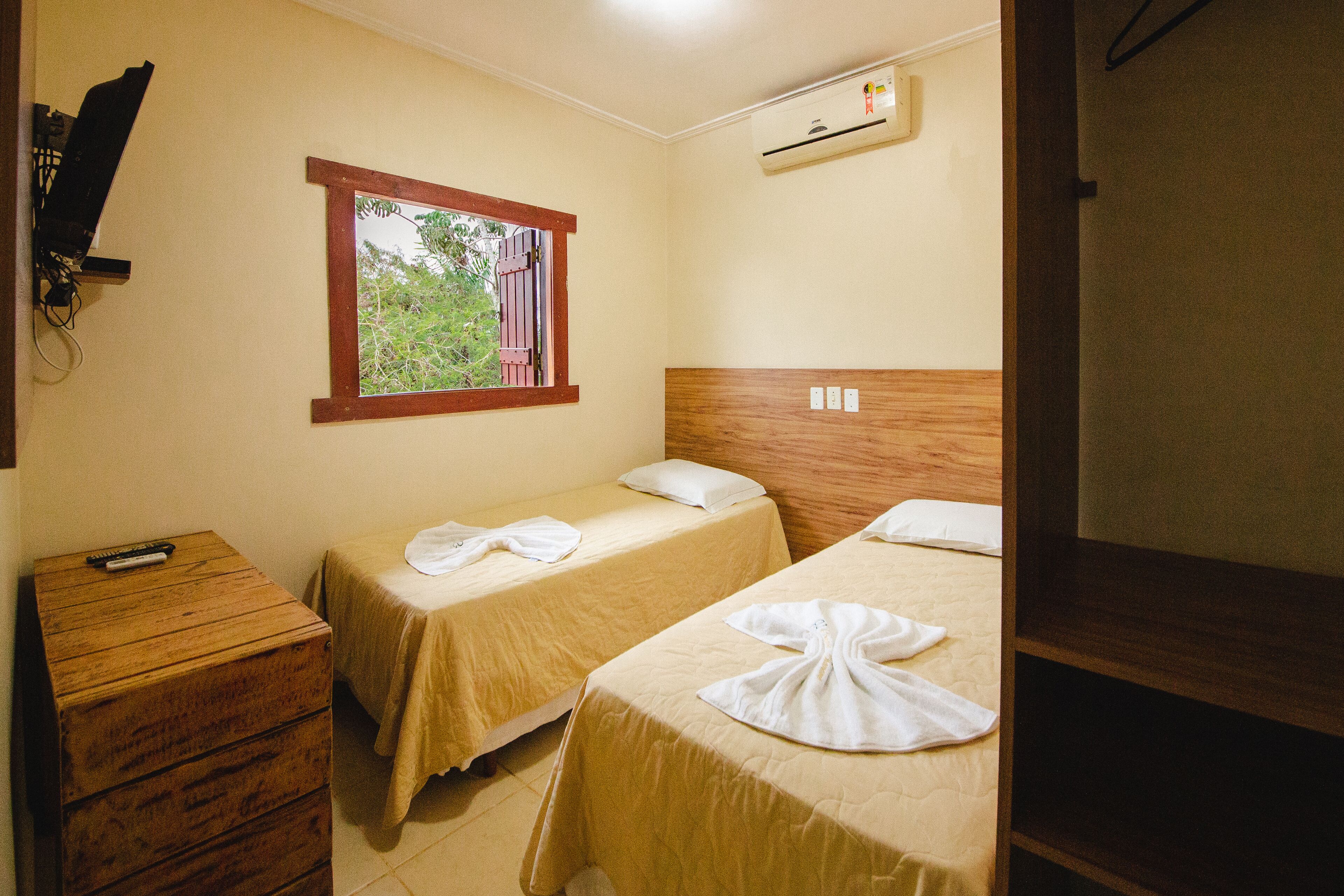 A hotel room with two single beds, a wooden dresser, a window with a view, and an air conditioning unit.