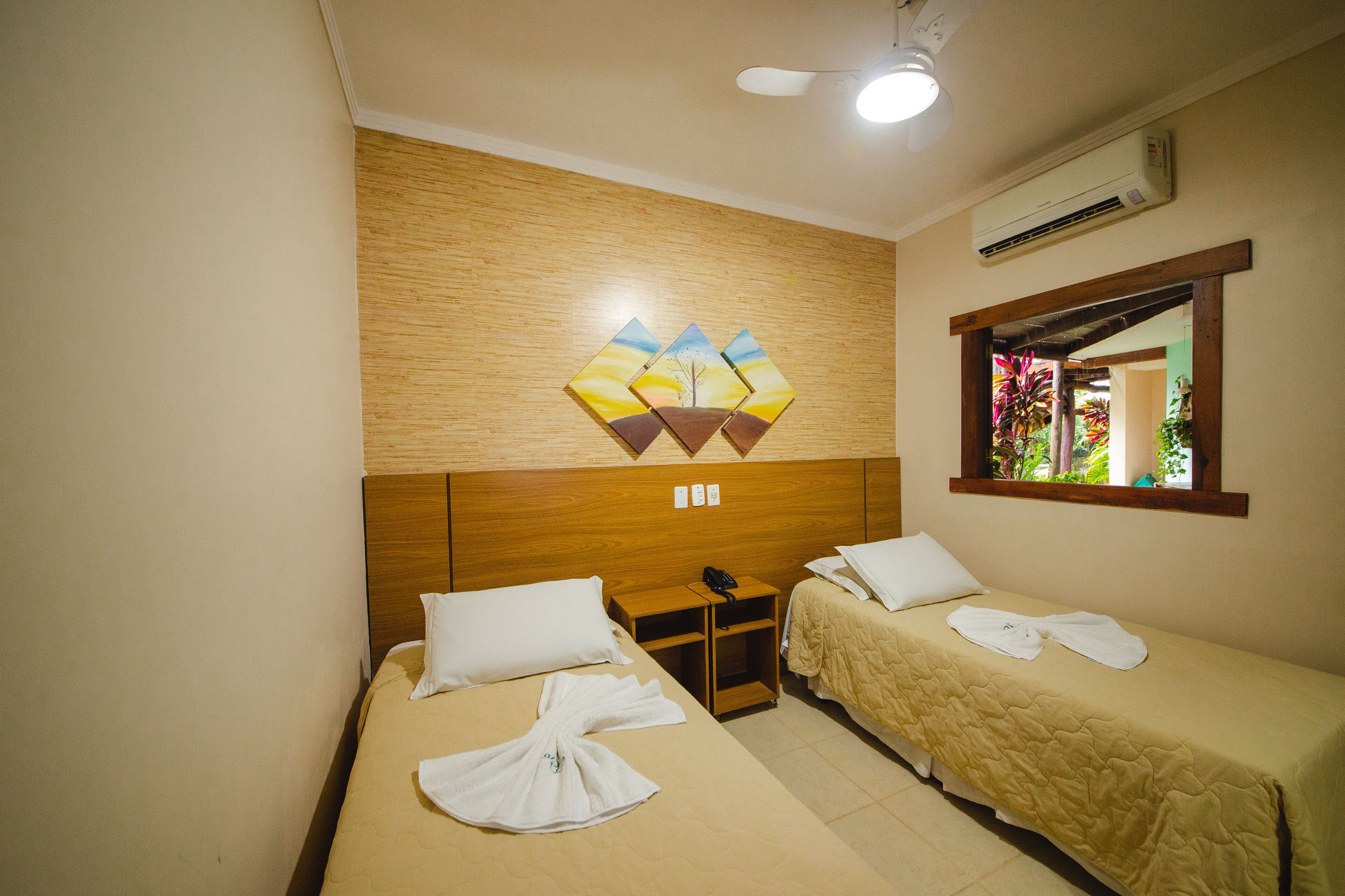 Una habitación de hotel con dos camas, cabecera de madera, aire acondicionado instalado en la pared y ventana con vistas a la vegetación.