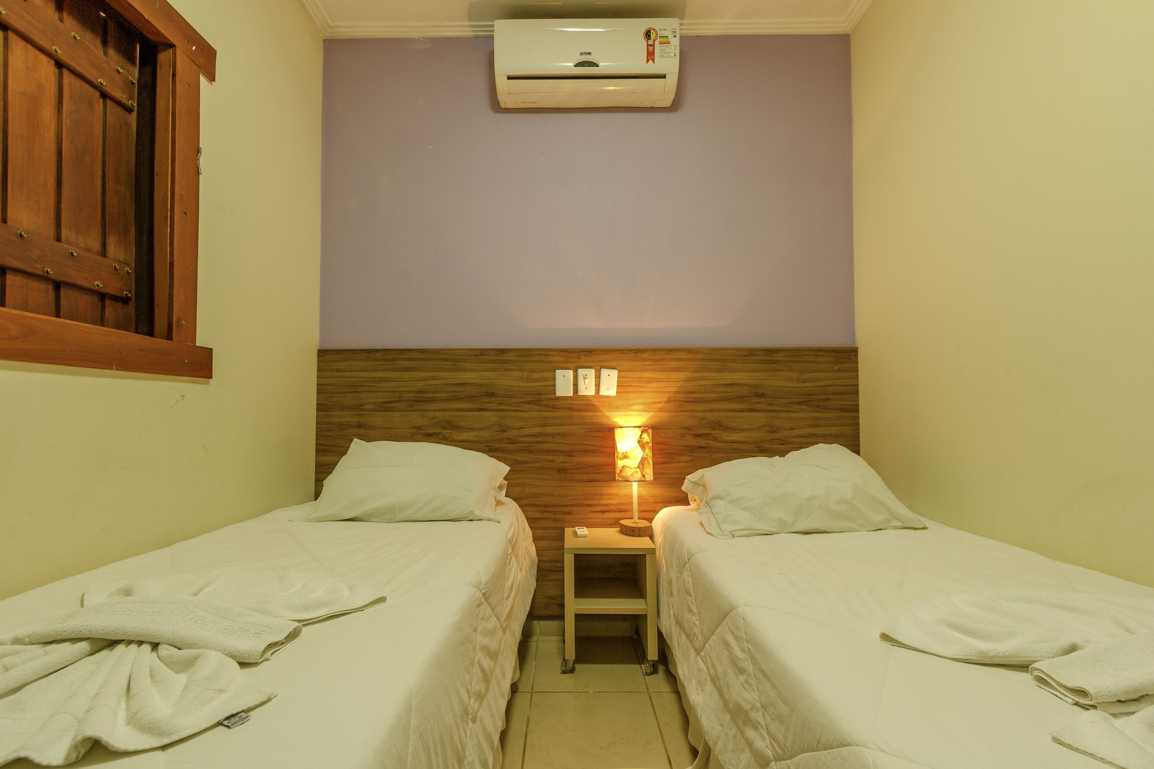 Habitación doble con dos camas, cabecera de madera, mesita de noche con lámpara y aire acondicionado en la pared.
