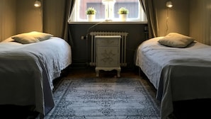 Una habitación con dos camas, un radiador, una mesa con dos plantas en macetas y una ventana con cortinas.