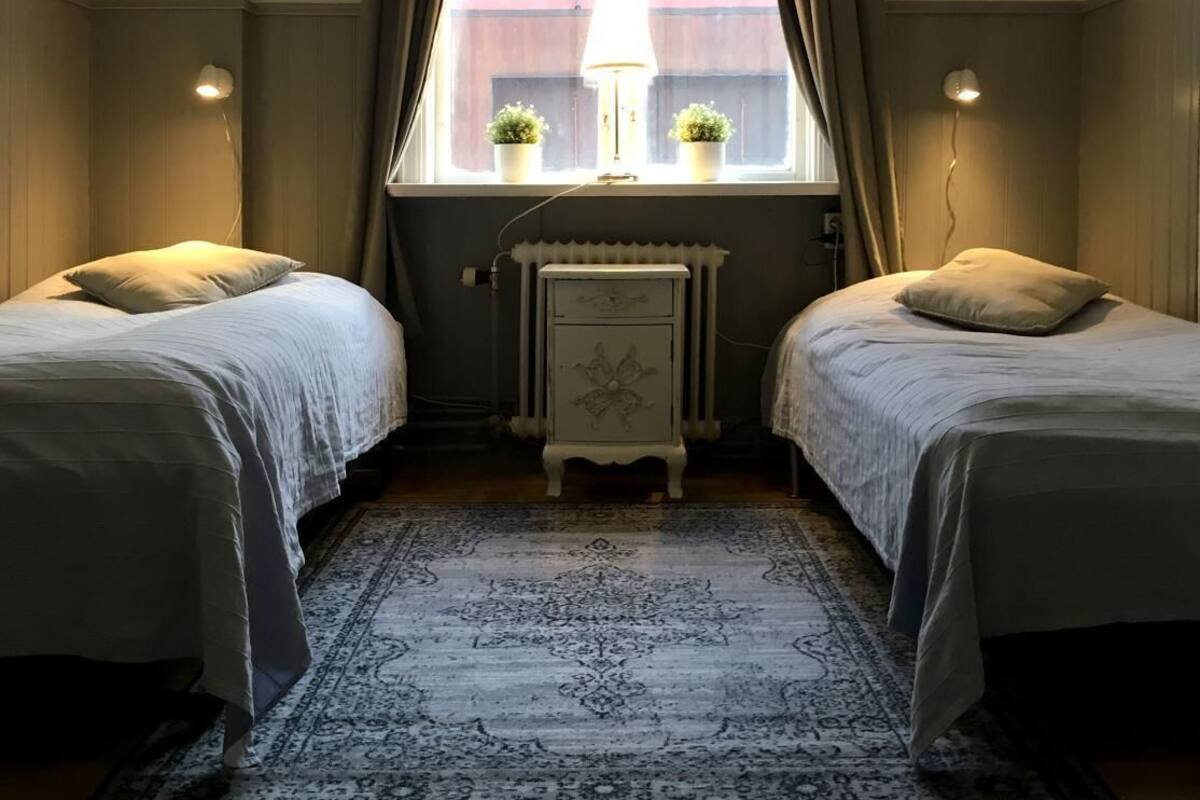 Een kamer met twee bedden, een radiator, een tafel met twee planten in potten, en een raam met gordijnen.