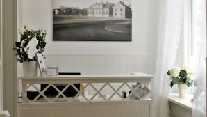 Una habitación con un mostrador de recepción blanco, una fotografía en blanco y negro enmarcada en la pared, una planta en maceta y una ventana con cortinas blancas.