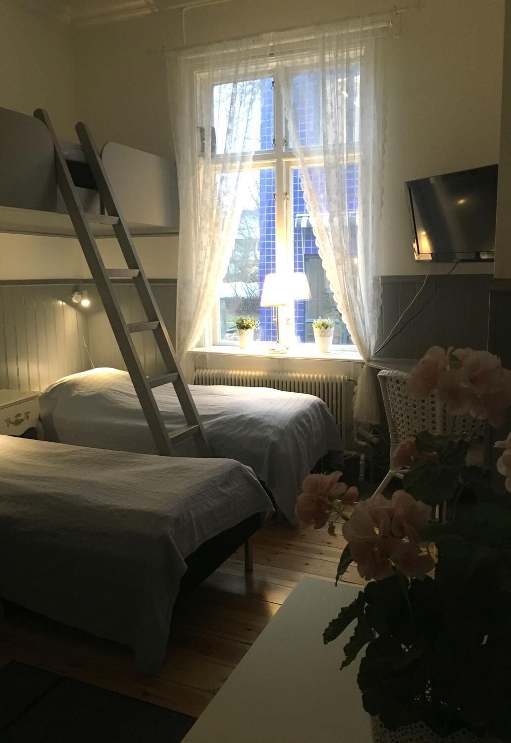 A room with two beds, a ladder, a radiator, a TV, and a window with curtains.