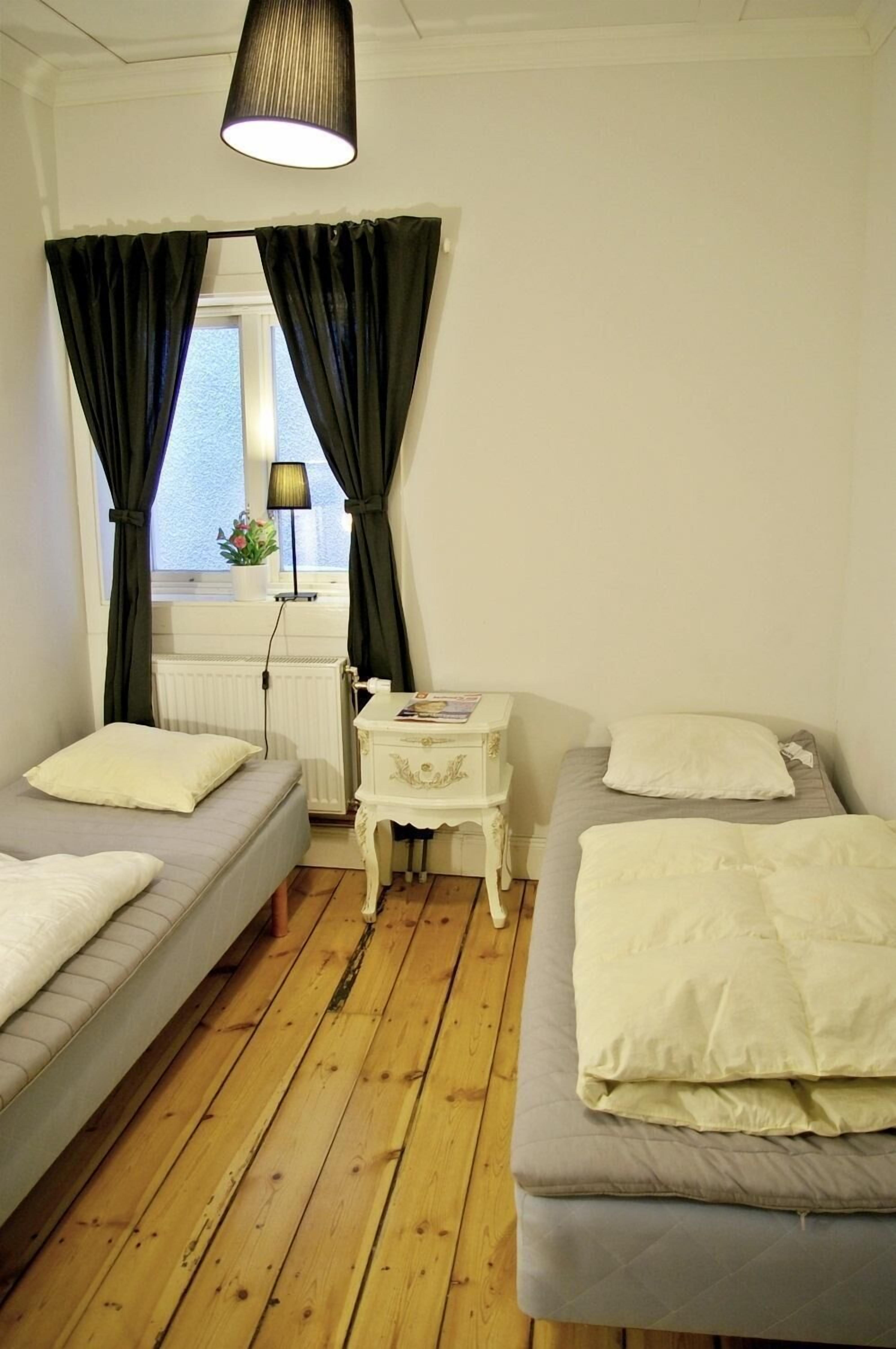 A room with two beds, a small table, a lamp, and a window with curtains.