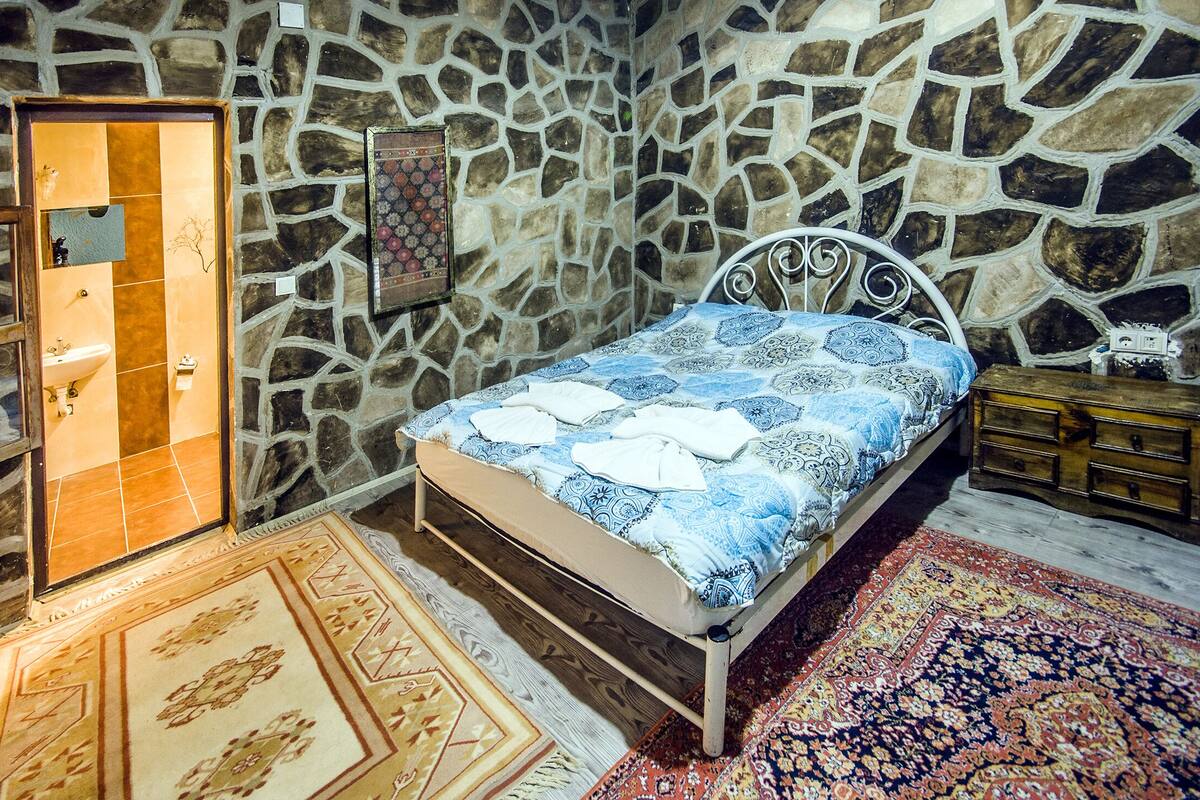 A bedroom with a stone wall, a bed with patterned bedding, a wooden nightstand, and a nightstand lamp.