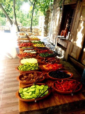 A variety of food items displayed on a wooden table outdoors, with a rustic stone wall and greenery in the background. - Kapadokya Organik Ciftlik Evi (Avanos)