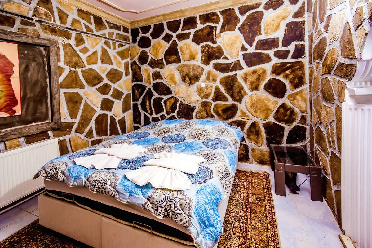 A room with a stone wall, a bed with patterned bedding, a framed picture, and a radiator.