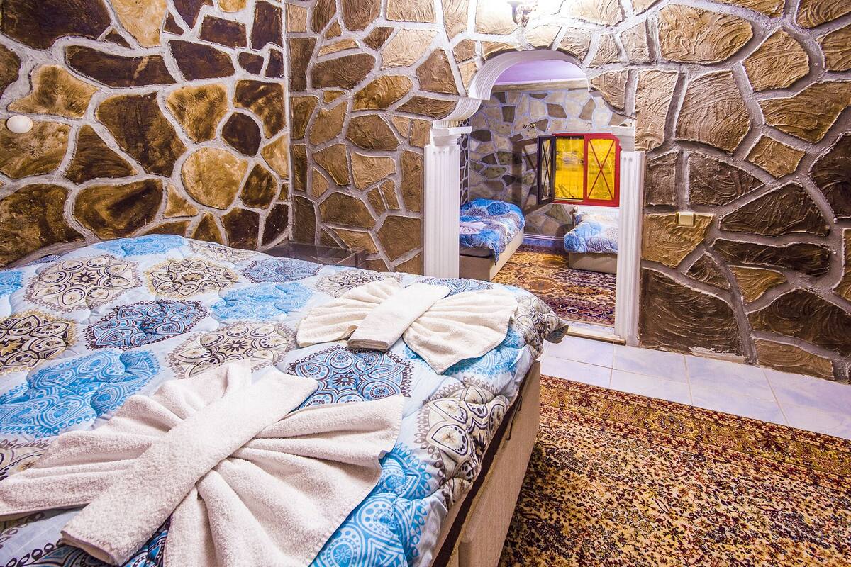 A stone-walled room with a bed, a small table, and a window with a yellow frame.