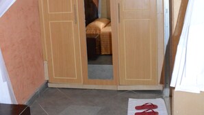 Un dormitorio con un armario de madera, una cama con cubrecama a rayas y un par de pantuflas rojas sobre una toalla en el suelo.