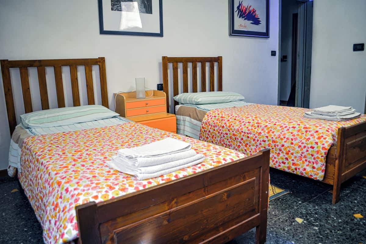 Une chambre avec deux lits en bois, chacun recouvert d’une courtepointe à motifs floraux et surmonté d’une serviette pliée. On y trouve une commode en bois avec une lampe, ainsi qu’un tableau encadré accroché au mur.